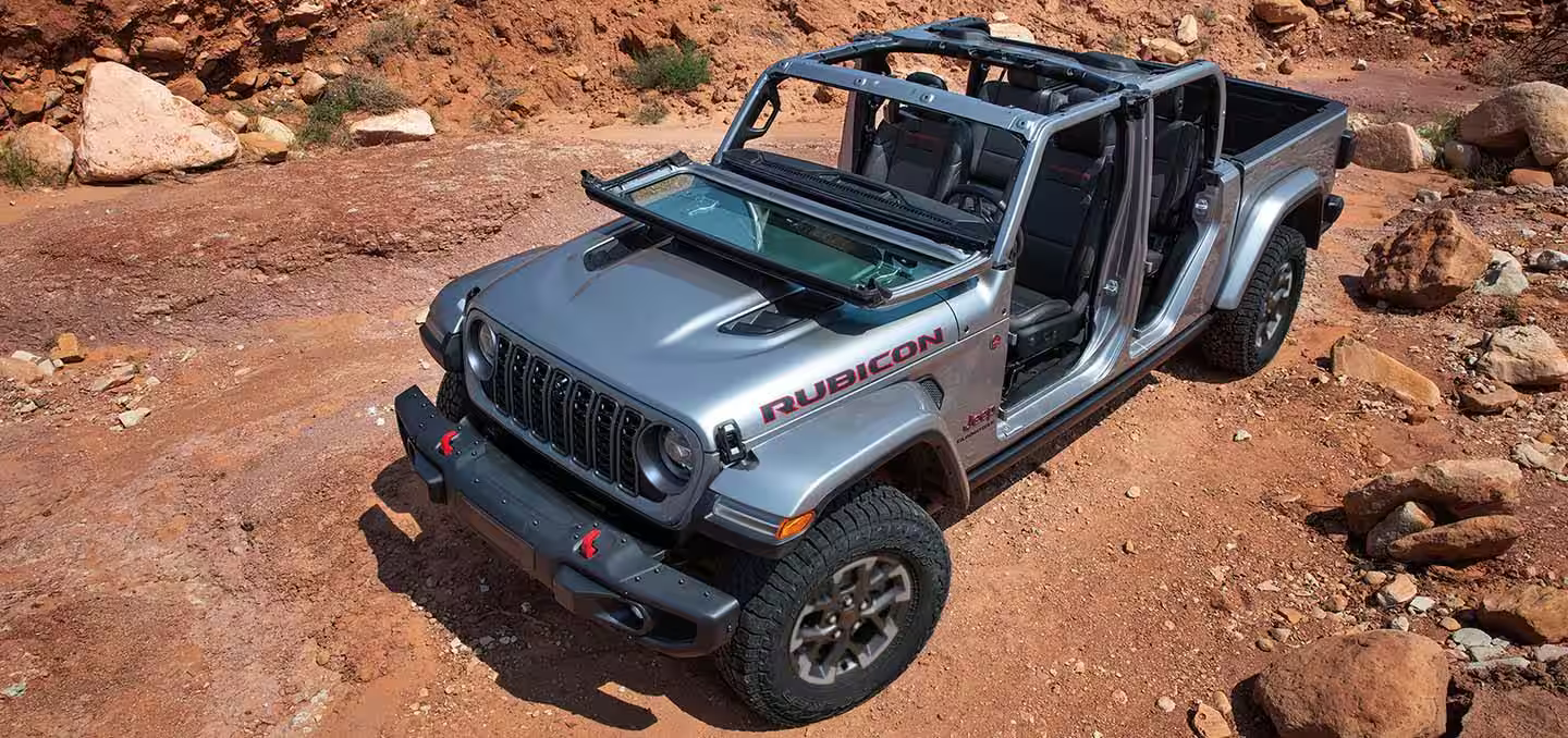 A silver 2024 Jeep Gladiator Rubicon with its windshield folded down and doors and roof removed, parked off-road in a clearing.