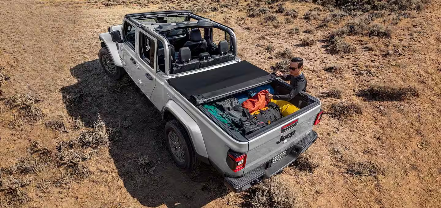A silver 2024 Jeep Gladiator Sport S with its roof removed and tonneau cover partially rolled up to reveal the truck bed packed tightly with camping gear.