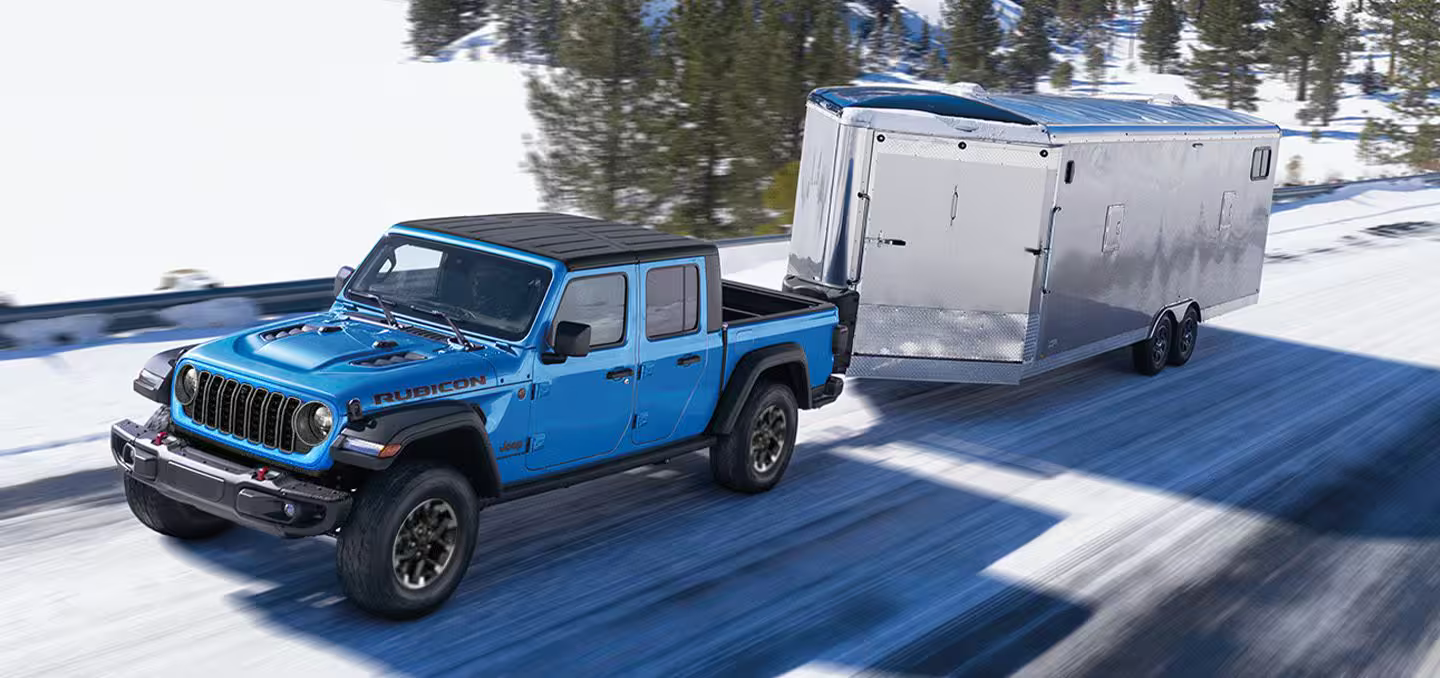 A blue 2024 Jeep Gladiator Rubicon with a black roof, being driven down a snow-covered highway as it tows an enclosed trailer.