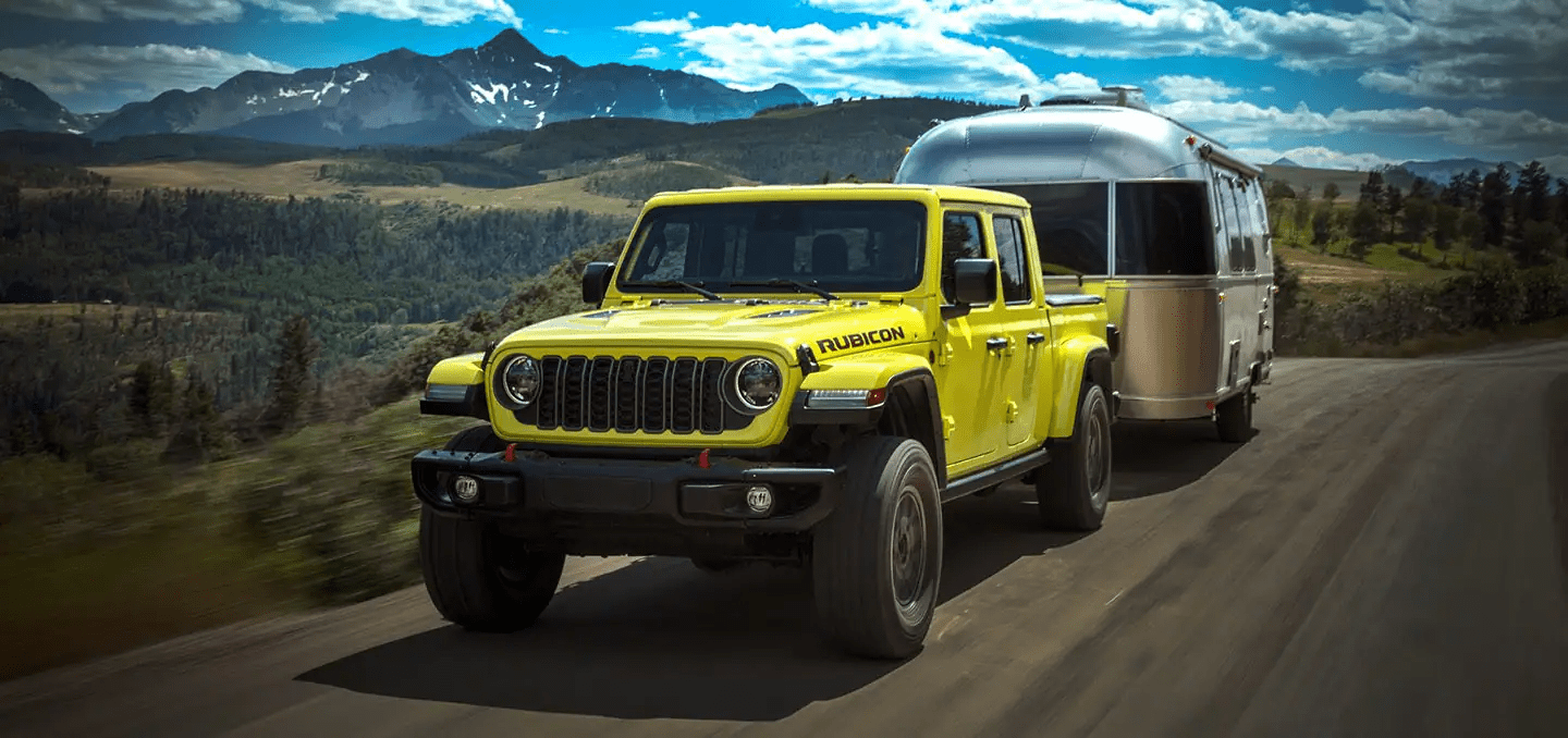A yellow 2024 Jeep Gladiator Rubicon being driven down a highway, towing a travel trailer.