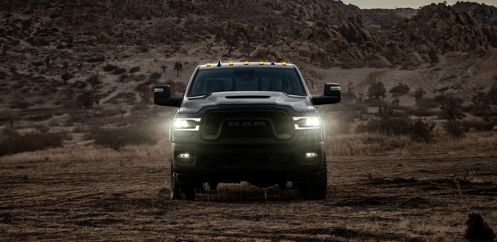 A head-on view of a RAM 2500 Rebel with its headlamps on, being driven off-road in the desert at dusk.